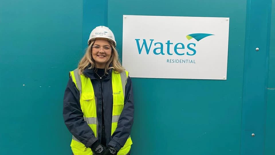 Alice is wearing a hi vis jacket and hard hat. She is smiling in front of a turquoise background with a white sign which has the Wates Residential logo on it.